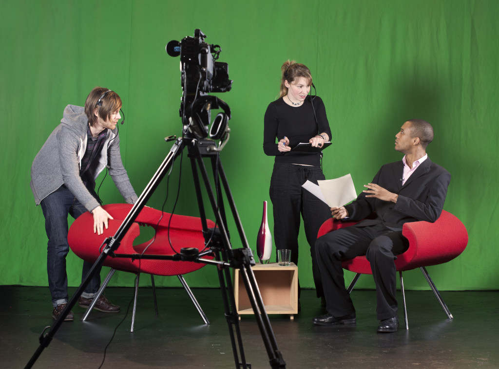 A crew member arranges furniture whilst the floor manager talks to the presenter on a TV studio set. TV camera on a tripod out-of-focus in the foreground.